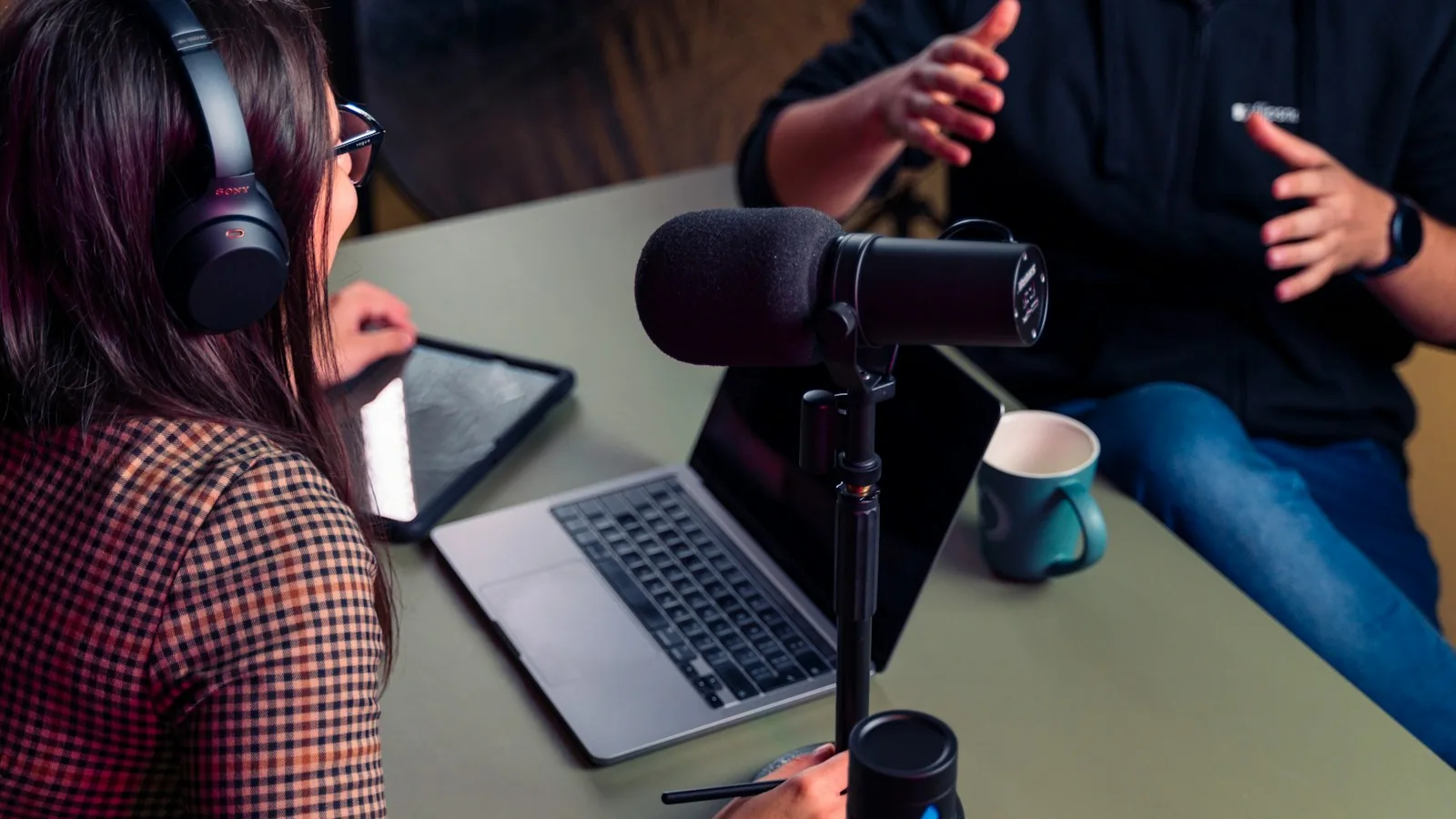 Two people wearing headphones having a conversation with a MacBook and podcast microphones on the desk