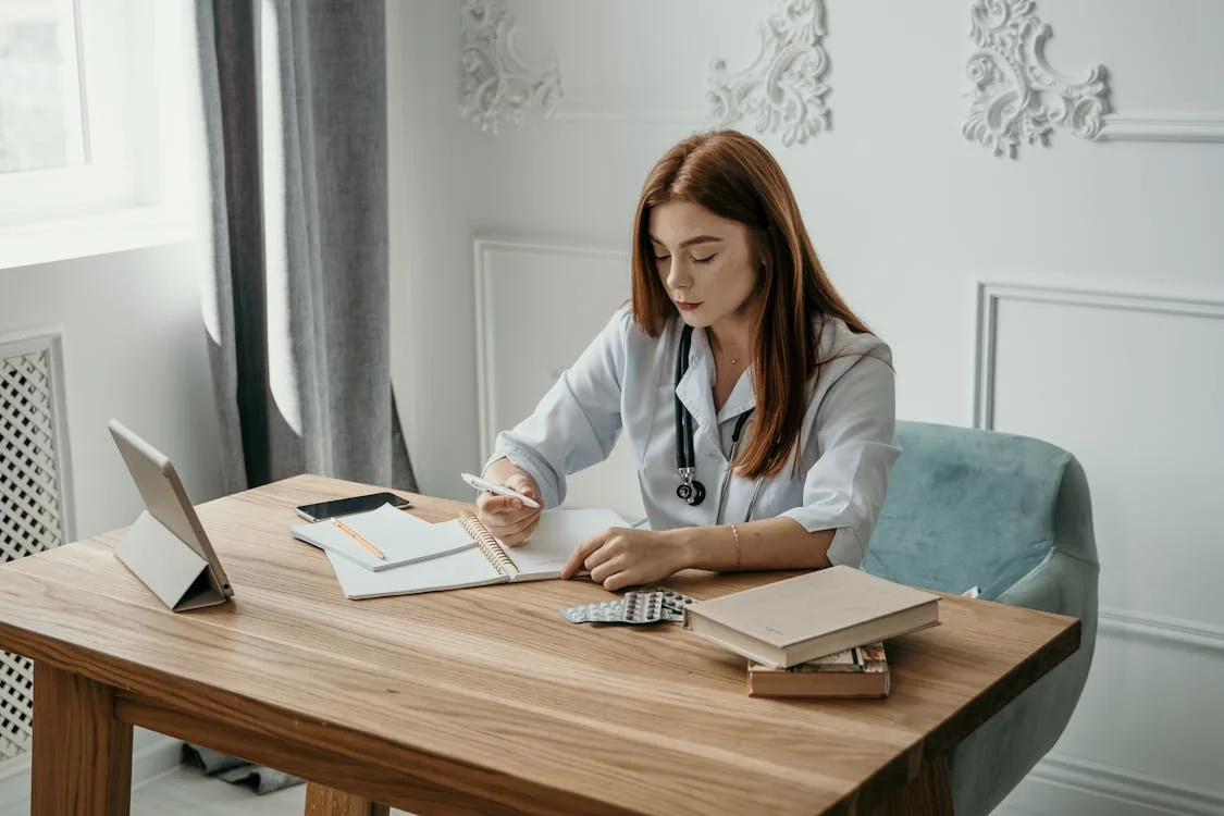 Female doctor at desk writing clinical notes with tablet and notebook — the documentation burden voice dictation solves